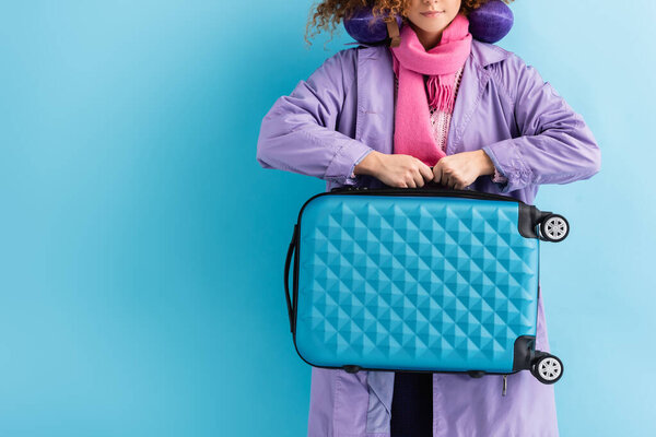 обрезанный вид молодой женщины в пальто и шарфе с чемоданом на синем