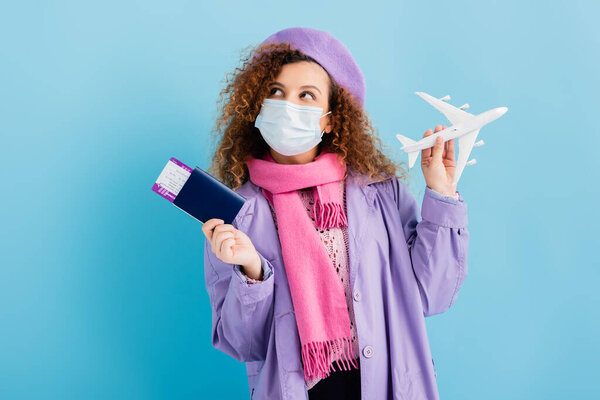 curly woman in beret, scarf, medical mask and coat holding passport and toy plane on blue