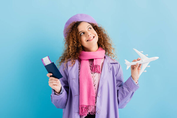 smiling woman in beret, scarf and coat holding passport and toy plane on blue