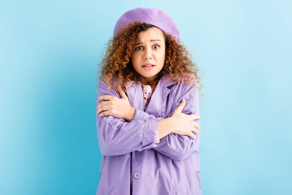curly woman in beret freezing and embracing herself on blue background 