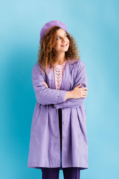 pleased young woman in beret and coat looking away on blue