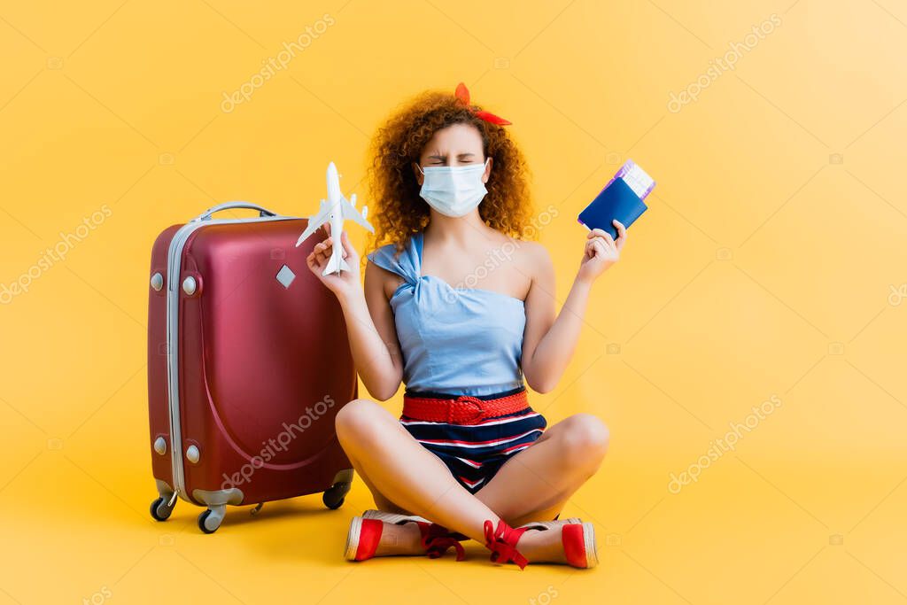 curly young woman in medical mask holding passport and toy plane while sitting near suitcase on yellow 