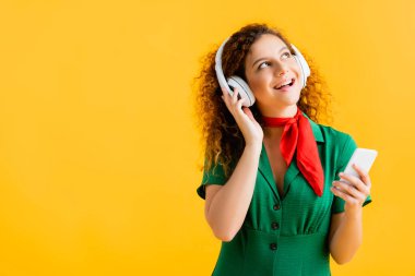 Kablosuz kulaklıklı neşeli kadın sarıda izole edilmiş akıllı telefon tutuyor. 