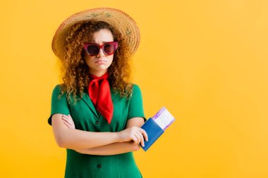 frustrated woman in straw hat, sunglasses and dress holding passport isolated on yellow  clipart