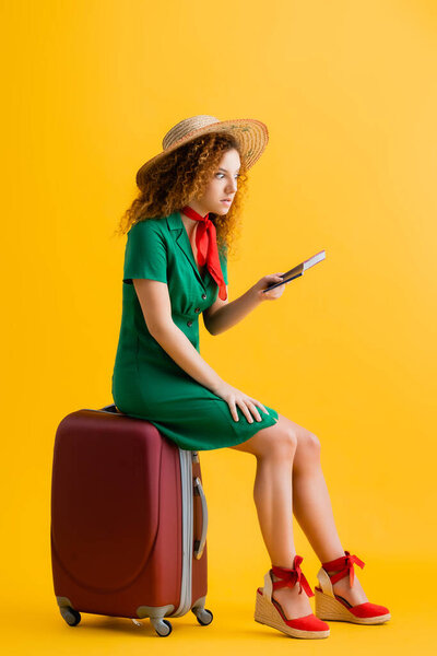 full length of angry woman in straw hat holding passport and sitting on luggage on yellow 