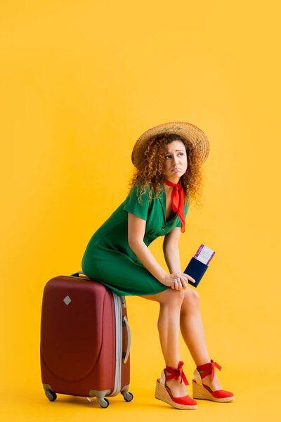full length of upset woman in straw hat holding passport and sitting on red luggage on yellow 