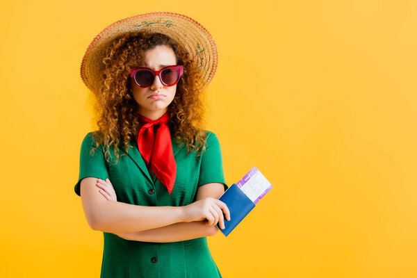 frustrated woman in straw hat, sunglasses and dress holding passport isolated on yellow 