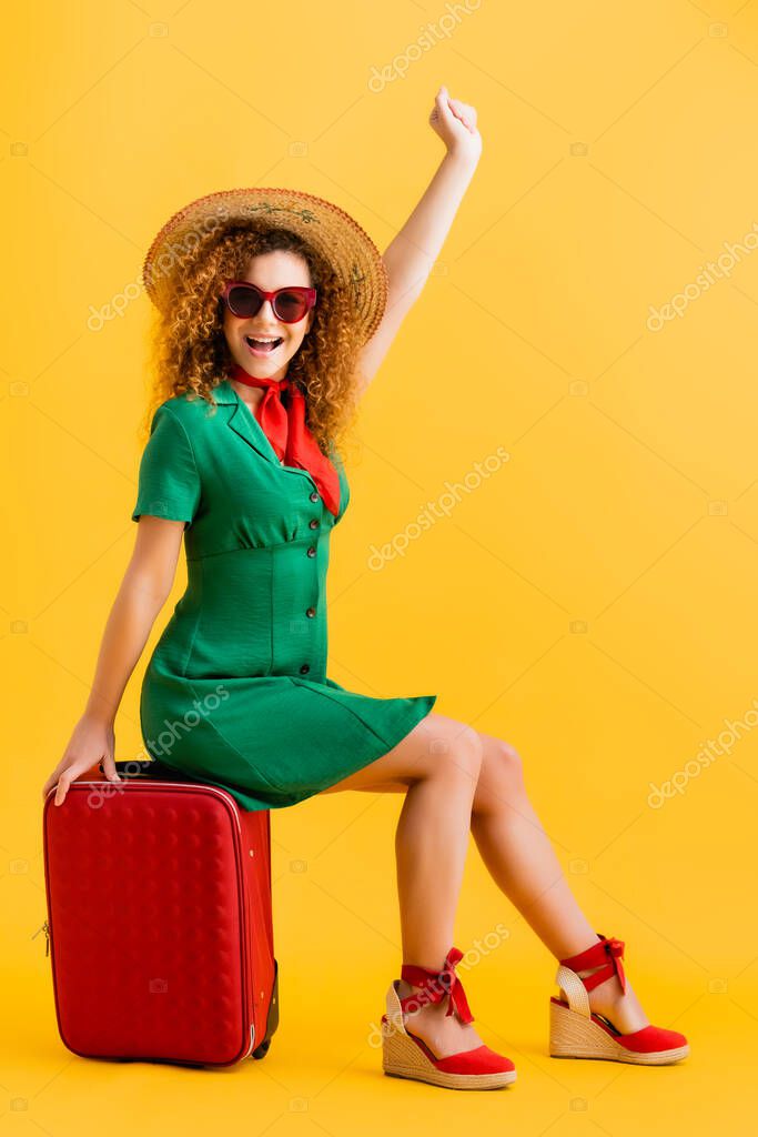 full length of excited woman in straw hat, sunglasses and dress sitting on luggage on yellow 