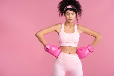 Spor giyim ve boks eldivenli kıvırcık bir kadın elleri pembe kalçalarında duruyor. 