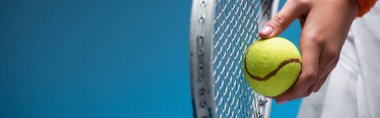 Mavi pankartta oynarken elinde tenis raketi ve top tutan genç bir kadın.