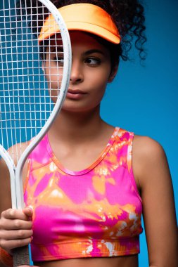 Sportif genç bir kadın elinde tenis raketi ile mavi renge bakarken gözlerini kaçırıyor.