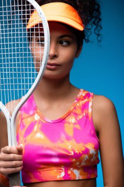 sportive young woman looking away while holding tennis racket on blue clipart