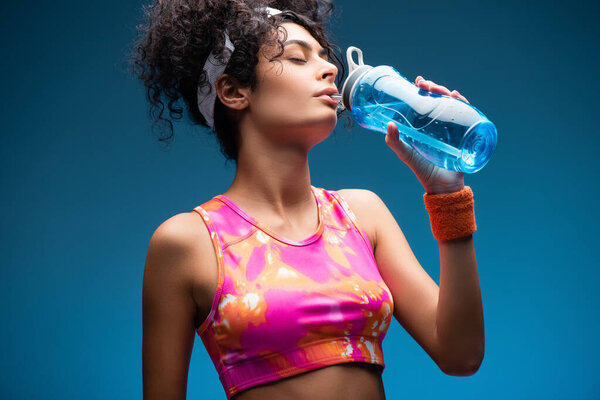 curly woman in sportswear drinking water while holding sports bottle on blue
