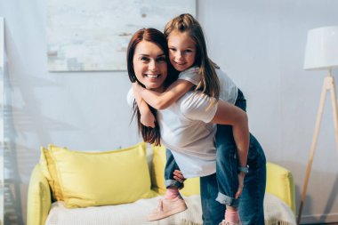 Neşeli anne, bulanık arka planda oturma odasında kızını sırtında taşırken kameraya bakıyor.