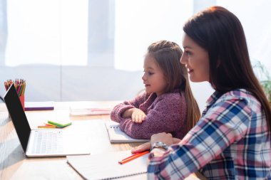 Gülümseyen anne ve kız masadaki laptopa bakıyor bulanık arka planda kırtasiyeyle.