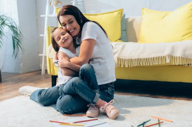 Mutlu, gözleri kapalı bir anne evde oturmuş kızını kucaklıyor.