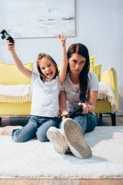 KYIV, UKRAINE - OCTOBER 19, 2020: Excited daughter with yes gesture holding joystick near sad mother on floor on blurred foreground clipart