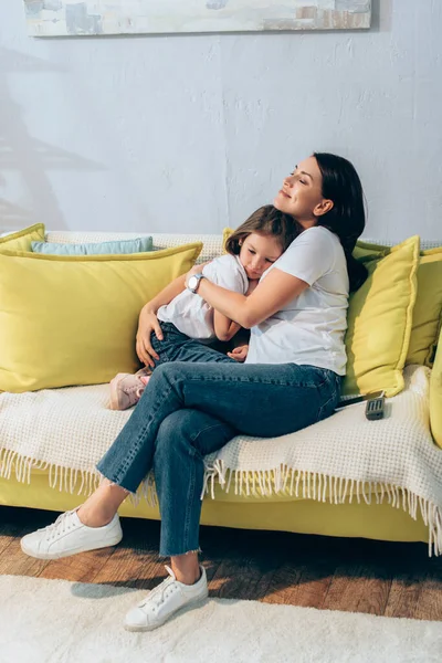 全长快乐的母亲 紧闭双眼 抱着女儿 坐在沙发上 — 图库照片