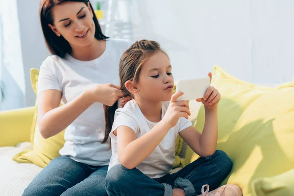 Leende Mor Flätning Hår Dotter Med Smartphone Suddig Bakgrund — Stockfoto