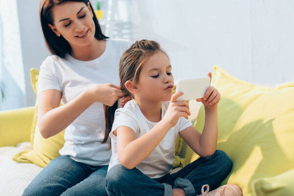 Smiling mother plaiting hair of daughter with smartphone on blurred background