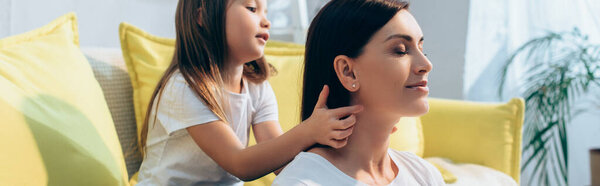 Daughter plaiting hair of smiling mother with closed eyes at home on blurred background, banner