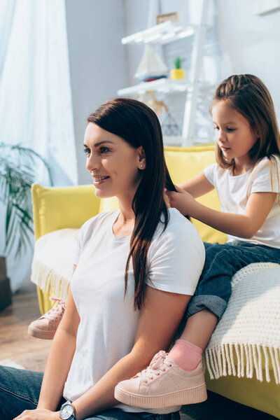 Daughter plaiting hair of smiling mother while sitting on couch at home on blurred background