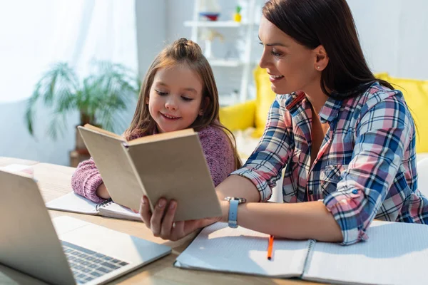 Smiling Mother Daughter Reading Book Desk Laptop Blurred Background — Stock Photo, Image