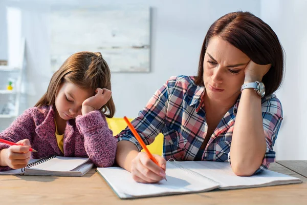 Erschöpfte Mutter Schreibt Kopierbuch Neben Tochter Schreibtisch Auf Verschwommenem Hintergrund — Stockfoto