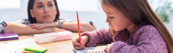 Волнуется мать, глядя на дочь, пишущую в блокноте за столом на размытом фоне, баннер