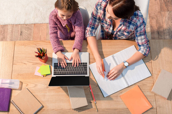 Вид сверху матери, сидящей рядом с дочерью и сидящей на ноутбуке за столом с канцелярскими принадлежностями дома