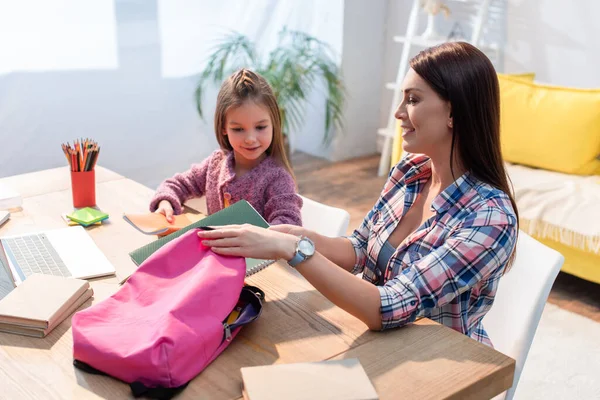 Happy Mother Putting Copy Book Backpack While Sitting Daughter Desk — Stock Photo, Image
