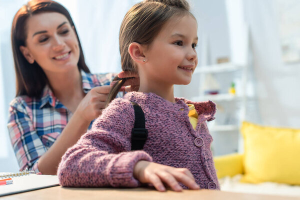 Happy mother plaiting hair of daughter with backpack at home on blurred background