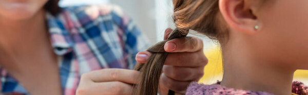 Cropped view of mother plaiting hair of daughter on blurred background, banner