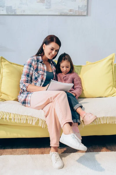 Full Length Smiling Mother Looking Copy Book Daughter Couch Home — Stock Photo, Image