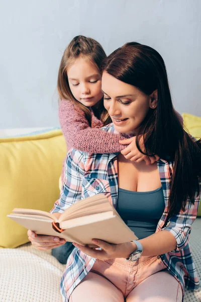 Daughter Looking Book While Hugging Smiling Mother Couch Home — Stock Photo, Image