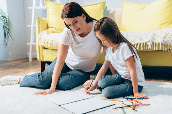 Mother sitting near daughter drawing in copy book on floor on blurred background