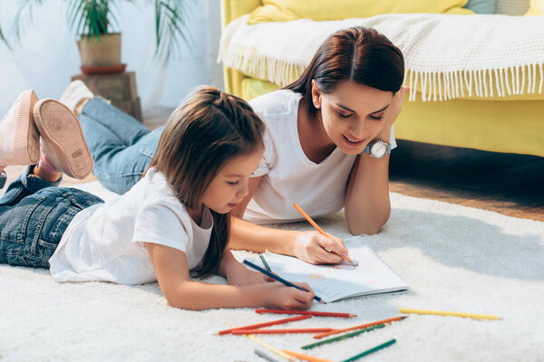 Positive mother and daughter with colored pencils drawing in copy book while lying on floor on blurred background