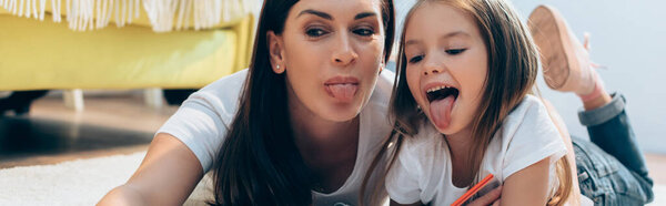 Happy mother and daughter with sticking out tongues looking down on floor on blurred background, banner