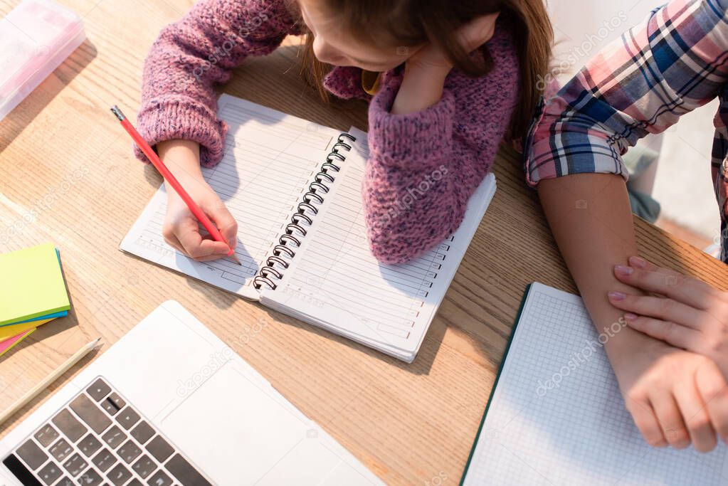 Top view of daughter writing in notebook near mother at desk with stationery and laptop