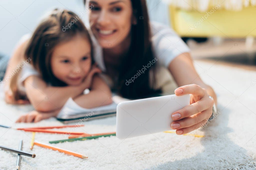 Cheerful mother and daughter taking selfie on floor on blurred background