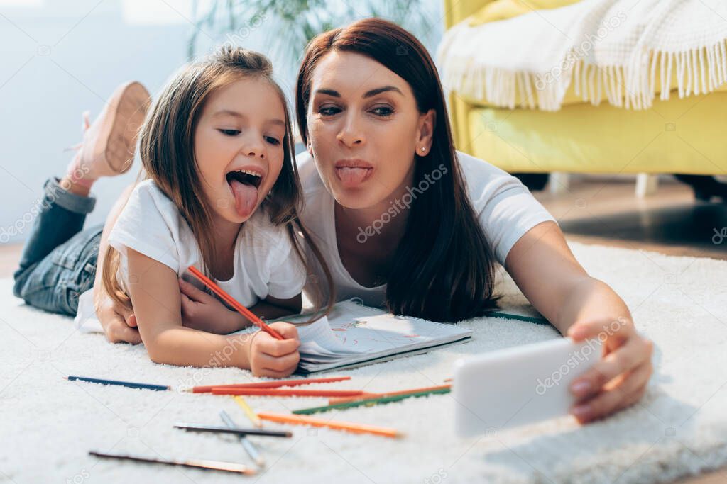 Happy mother and daughter with sticking out tongues taking selfie while lying on floor with colored pencils on blurred foreground