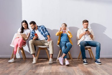 Çok kültürlü insanlar ve hamile bir kadın akıllı telefonlar kullanıyor ve kuyruktaki sandalyeler üzerinde kitap okuyor. 