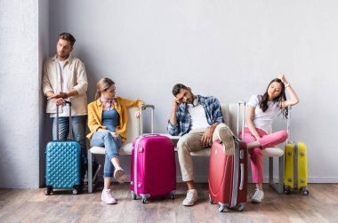 Tükenmiş çok kültürlü insanlar havaalanındaki koltuklarda bavullarının yanında oturuyorlar. 