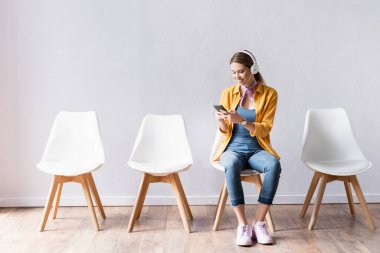 Koridorda akıllı telefon kullanan kulaklıklı, gülümseyen bir kadın. 