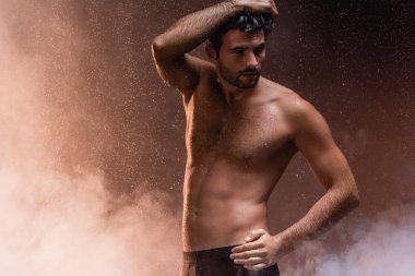 Seksi gömleksiz adam, eli kalçasında, yağmurun altında karanlık arka planda dumanla poz veriyor.