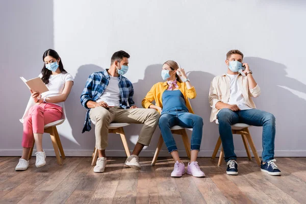 Pessoas Multiculturais Máscaras Médicas Falando Lendo Livro Sobre Cadeiras Salão — Fotografia de Stock