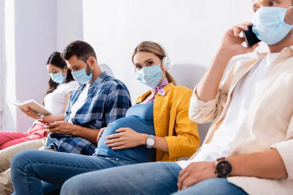 Pregnant woman in medical mask and headphones sitting near multiethnic people on blurred background in hall