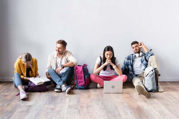 Multicultural students with laptop and book sitting on floor in hall 