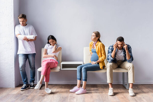 Мультиэтнические люди стоят в очереди рядом с беременной женщиной и мужчиной с гипсом под рукой в клинике 