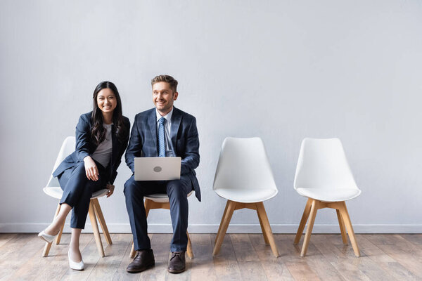 Smiling multicultural business people with laptop looking at camera on chairs in hall 